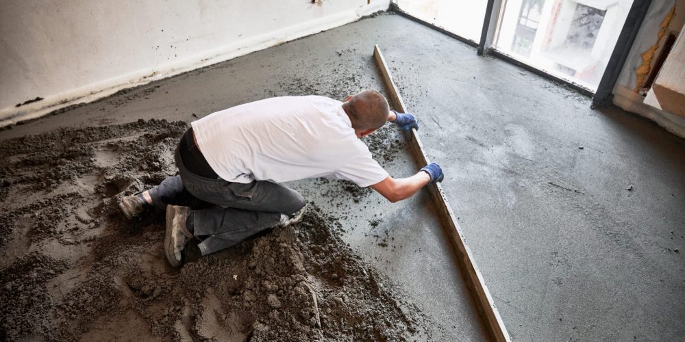 Male construction worker placing screed rail on the floor covered with sand-cement mix. Man smoothing and leveling surface with straight edge while screeding floor in apartment.
